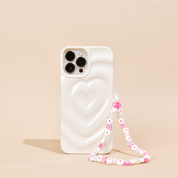 A white smartphone with a heart-shaped design on its back cover stands upright against a beige background. Attached is the Beaded Phone Strap - Flowers & Pearls by Coconut Lane, featuring pink and white beads, including charming flower-shaped ones, making it the perfect decorative accessory.