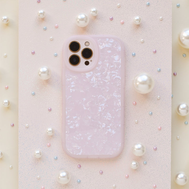 A smartphone encased in a Moonstone Pink Phone Case by Coconut Lane, glossy and textured, rests on a surface adorned with various sizes of pearls. The camera lenses are visible at the top left of the phone. The background features a slightly glittery pastel finish, adding an elegant touch.