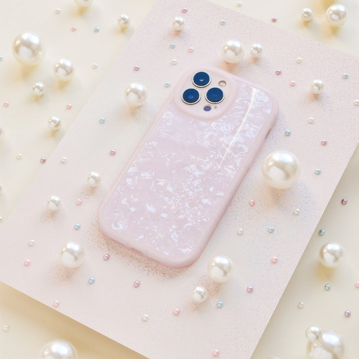 A Moonstone Pink Phone Case by Coconut Lane, featuring a pale pink hue with a pearlescent finish, is showcased on a textured pink surface. Variously sized white pearls and small multicolored beads encircle the case, creating an elegant and whimsical display.