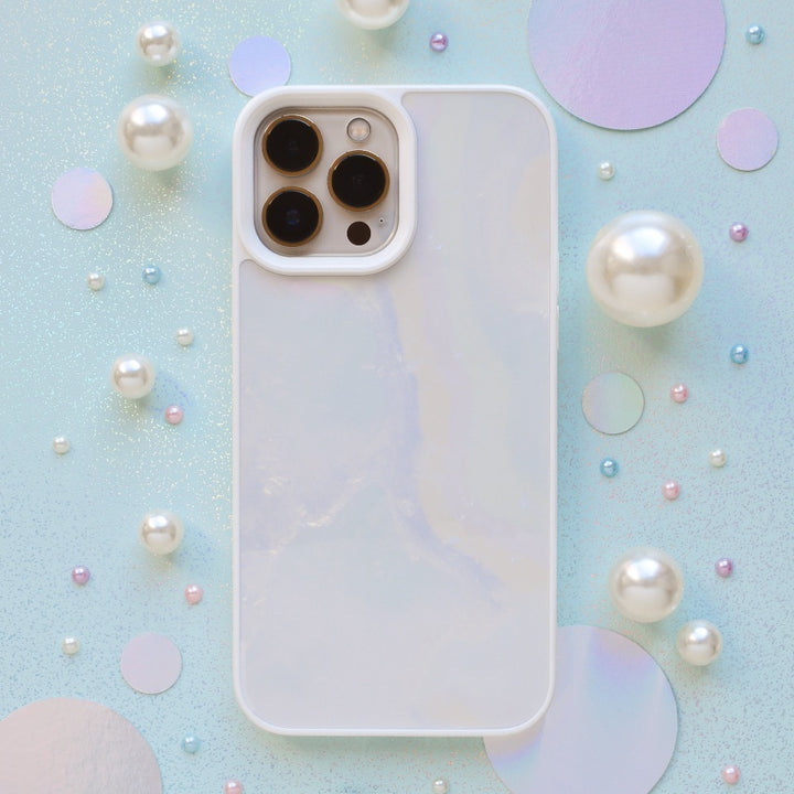 A white iPhone15 with a Coconut Lane Mother of Pearl Phone Case is placed on a pastel background adorned with scattered faux pearls and iridescent circular discs. The phone, showcasing its three camera lenses and sleek design, exhibits an elegant pearlescent effect.