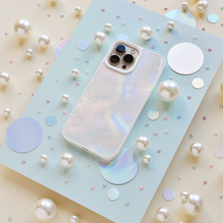 A Mother of Pearl Phone Case from Coconut Lane lies on a light blue, glittery surface surrounded by various sizes of white pearls and holographic circles. The pearlescent effect creates an aesthetic and dreamy visual effect.