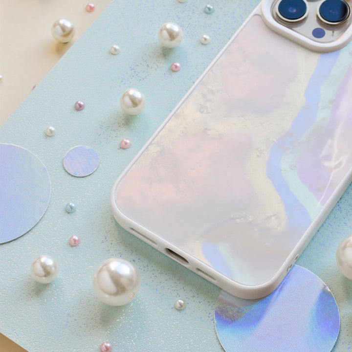 A pastel-colored smartphone with Coconut Lane's Mother of Pearl Phone Case lies on a light blue background adorned with various sizes of glossy white and pastel-colored beads and circular holographic cutouts.