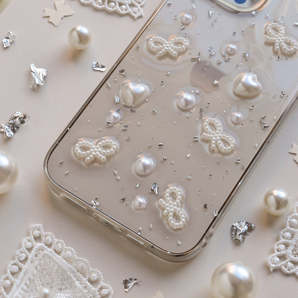 A close-up image of the Coquette Dreams Phone Case by Coconut Lane, a transparent iPhone 15 case adorned with 3D white butterfly and heart designs, as well as pearl-like embellishments. Extra protective, the phone rests on a white surface surrounded by decorative pearls, lace cutouts, and silver confetti pieces.