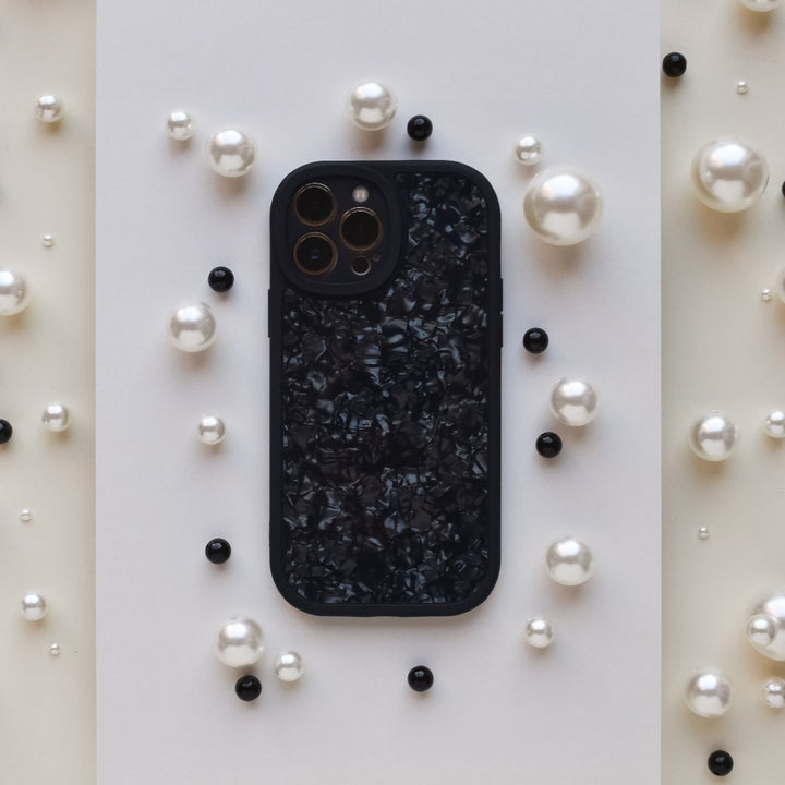 A Moonstone Black Phone Case by Coconut Lane, featuring a textured, glossy finish, is centered on a white rectangular background. The phone case is surrounded by scattered pearls in various sizes and a few black round beads, creating a contrasting and elegant arrangement that showcases its durable protection.
