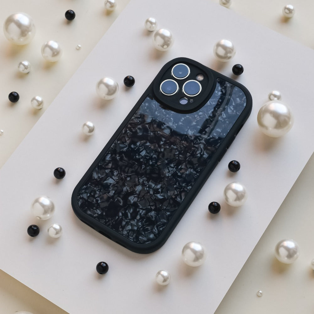 A smartphone with a dark, textured back cover lies on a white surface. Surrounding the phone are scattered white and black pearl-like beads, providing an elegant contrast to the sleek device, showcasing its sophisticated Moonstone Black Phone Case by Coconut Lane that offers durable protection.
