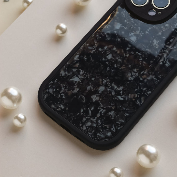 A Moonstone Black Phone Case by Coconut Lane with a pearlescent pattern lies on a flat surface surrounded by scattered pearls of various sizes. The phone case has a cutout for the camera module, which is partially visible at the top right corner, offering durable protection while maintaining elegance.