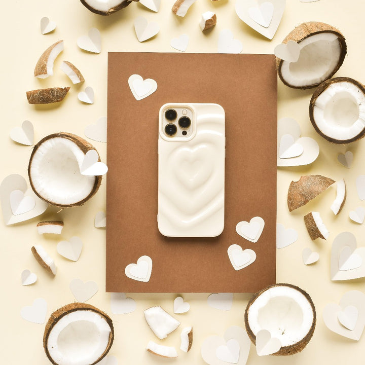 A white smartphone with the Melting Heart Phone Case - Coconut Cream by Coconut Lane lies on a brown rectangular surface. Surrounding the phone are halved coconuts and scattered coconut pieces mixed with white paper heart cutouts, all arranged on a beige background.