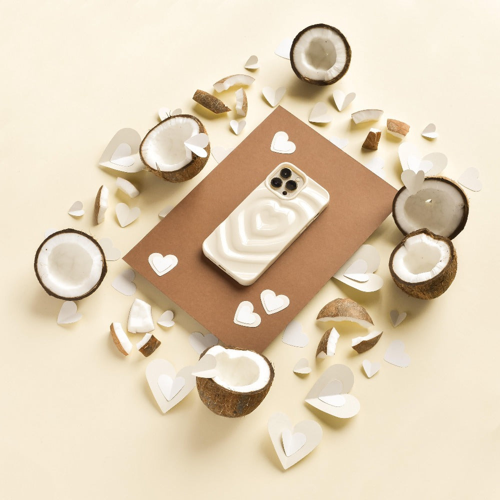 A smartphone with the Melting Heart Phone Case - Coconut Cream from Coconut Lane lies on a brown rectangular sheet. Surrounding it are halved coconuts and heart-shaped paper cutouts scattered on a cream-colored surface, creating a warm, tropical, and romantic ambiance.
