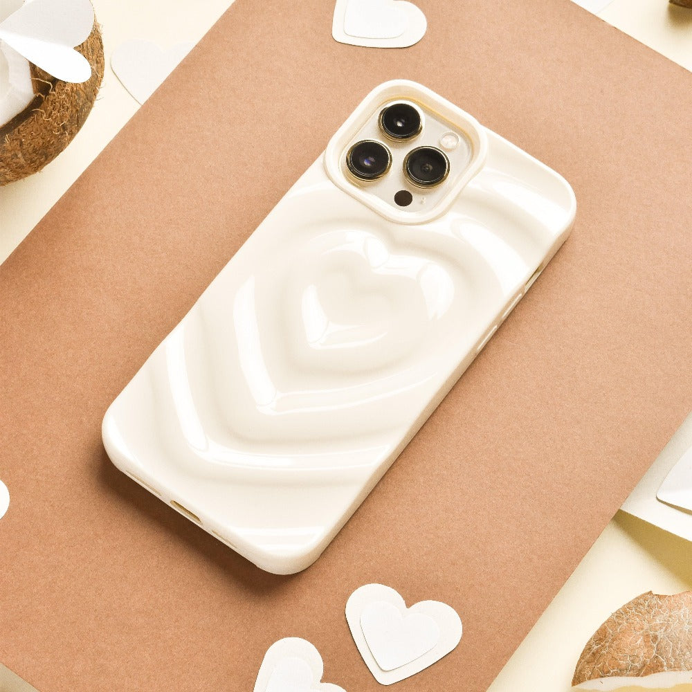 A Melting Heart Phone Case - Coconut Cream by Coconut Lane, with its smooth, pale beige case featuring a raised, 3D heart design, lies on a piece of brown paper. Surrounding the phone are small white heart-shaped cutouts and halved coconuts. The background showcases cream-colored and various brown tones.