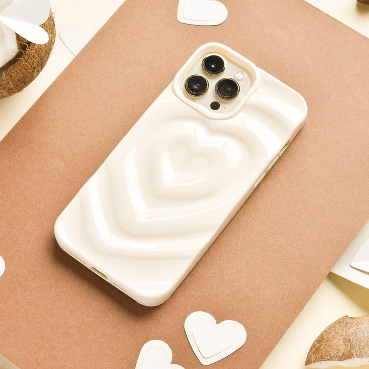 A Melting Heart Phone Case - Coconut Cream by Coconut Lane, with its smooth, pale beige case featuring a raised, 3D heart design, lies on a piece of brown paper. Surrounding the phone are small white heart-shaped cutouts and halved coconuts. The background showcases cream-colored and various brown tones.
