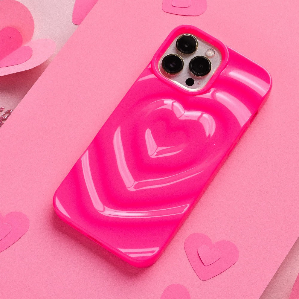 The Melting Heart Phone Case - Bright Pink by Coconut Lane, featuring a raised heart design with a 3D melting heart effect, is lying on a pink surface. The extra protective phone case is surrounded by small paper hearts, enhancing the overall pink-themed, romantic aesthetic.