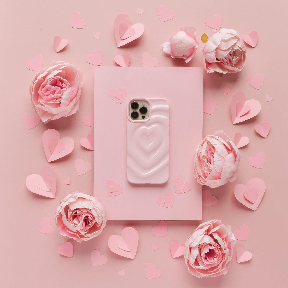 A pink smartphone adorned with Coconut Lane's Melting Heart Phone Case in Peony Pink is centered on a pink background. The phone is encircled by pink paper hearts of various sizes and clusters of light pink flowers, creating a romantic and delicate ambiance.