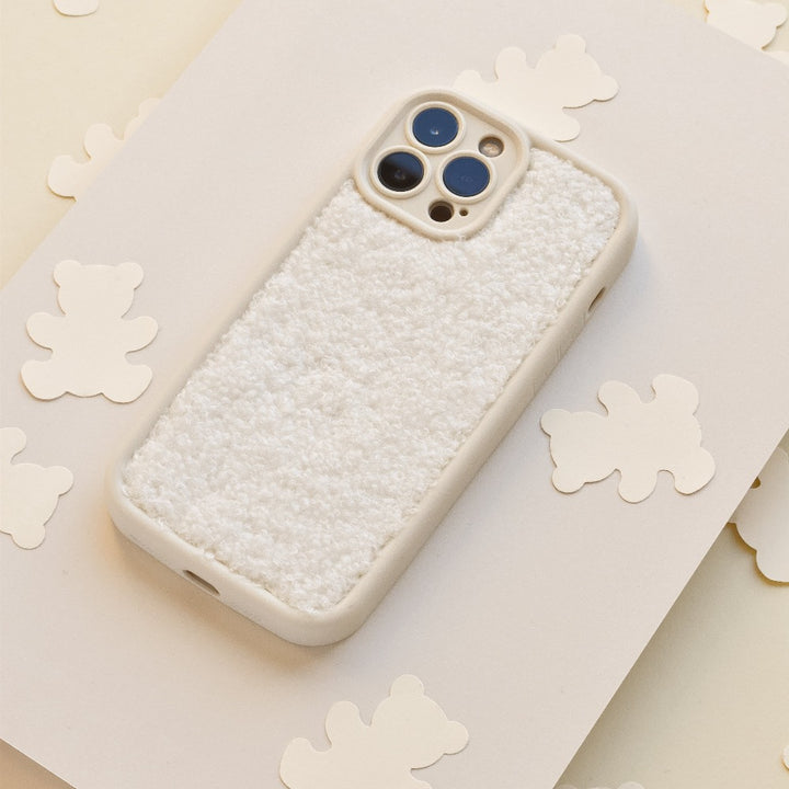 A **Cosy Teddy Phone Case - Cream** by *Coconut Lane*, designed for a smartphone with three camera lenses on the back, lies on a beige surface. The extra protective case features a fuzzy texture adorned with small white teddy bear-shaped cutouts scattered around it.