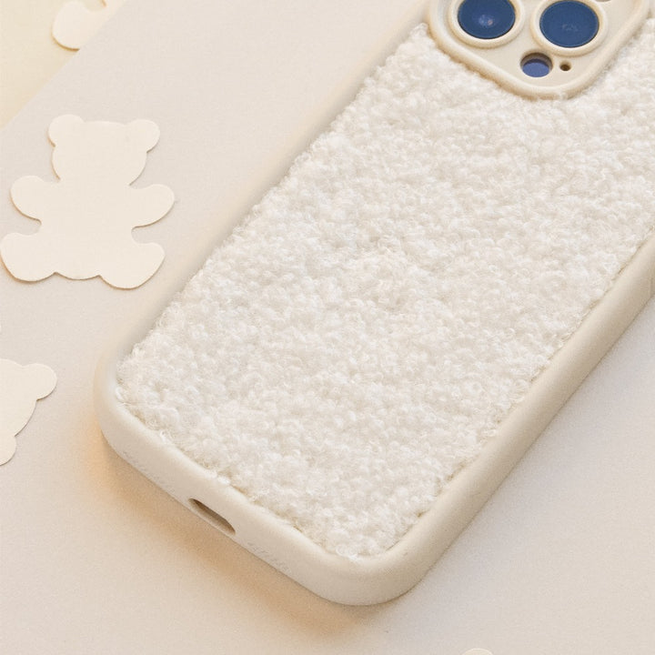 A smartphone rests on a surface with a Cosy Teddy Phone Case - Cream by Coconut Lane, designed to resemble soft fabric. The camera lens is exposed, and the plush material of the case looks extra protective. Surrounding the phone are cream teddy bear-shaped cutouts, adding a playful touch to the scene.