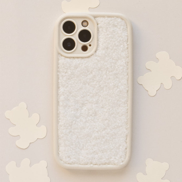 A Cosy Teddy Phone Case - Cream by Coconut Lane, featuring a textured teddy finish and light color, is placed on a flat surface surrounded by small bear-shaped cutouts. The case, which has openings for the camera lenses, appears soft to touch.