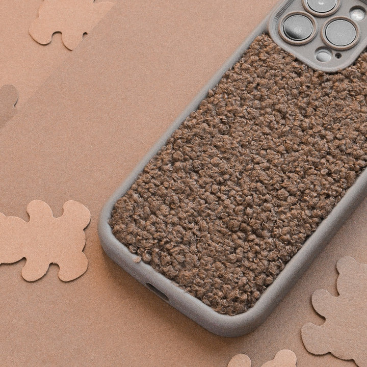 A close-up photo of a smartphone with a textured, brown, and rugged Cosy Teddy Phone Case - Brown by Coconut Lane resting on a surface scattered with various brown shapes. The phone's camera lenses are prominently visible at the top right corner.