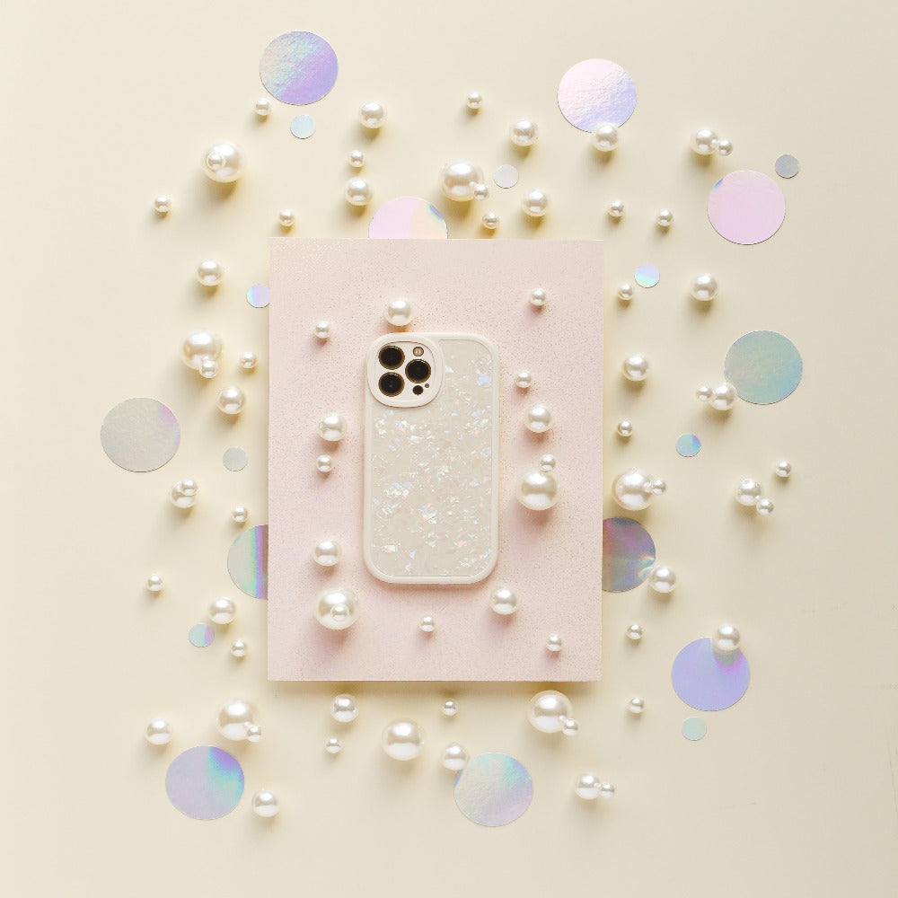A smartphone in the Moonstone Pearl Phone Case by Coconut Lane, featuring a sparkly, translucent design, is placed on a light pink rectangular paper. Surrounding it are scattered white pearls and iridescent circular confetti on a light beige background, creating a decorative and elegant arrangement.
