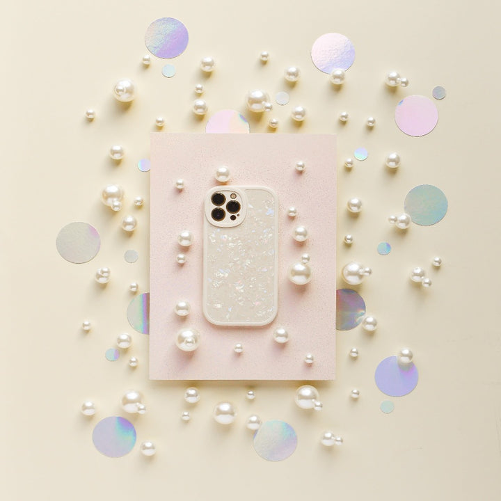 A smartphone in the Moonstone Pearl Phone Case by Coconut Lane, featuring a sparkly, translucent design, is placed on a light pink rectangular paper. Surrounding it are scattered white pearls and iridescent circular confetti on a light beige background, creating a decorative and elegant arrangement.