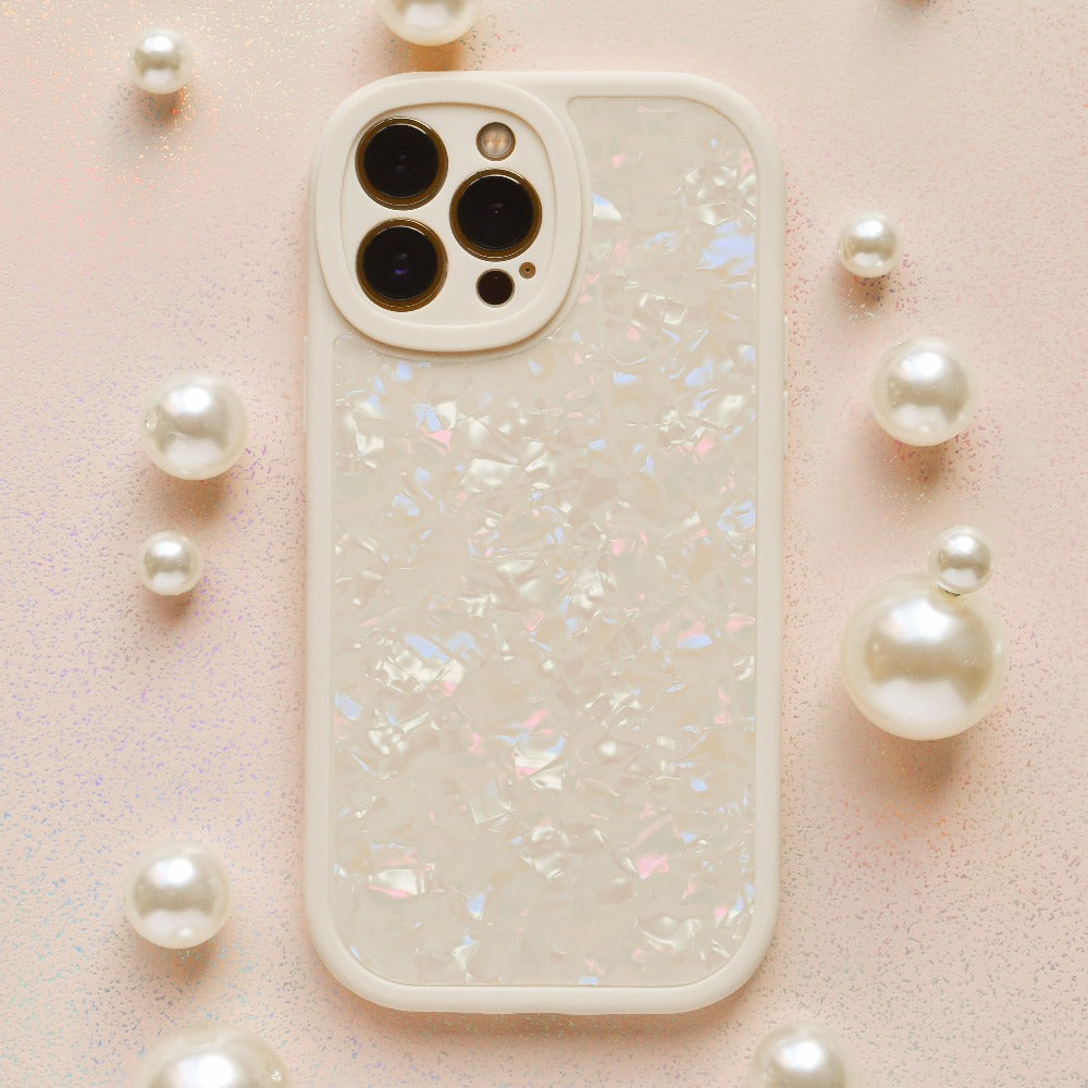 A smartphone protected by the Coconut Lane Moonstone Pearl Phone Case, showcasing an iridescent shell design, is placed on a light pink glittery surface. The phone, potentially an iPhone15 case model, features three rear cameras and is encircled by small and large pearl-like decorative spheres.