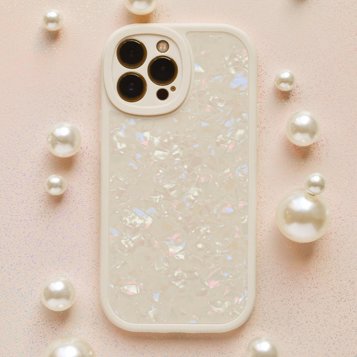 A smartphone protected by the Coconut Lane Moonstone Pearl Phone Case, showcasing an iridescent shell design, is placed on a light pink glittery surface. The phone, potentially an iPhone15 case model, features three rear cameras and is encircled by small and large pearl-like decorative spheres.