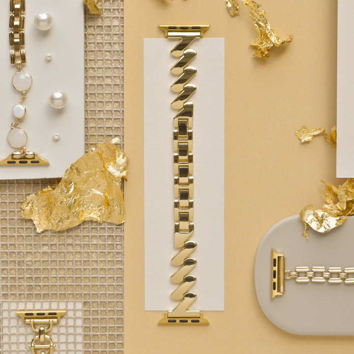A photo showcasing the Get Wavy Apple Watch Strap - Gold by Coconut Lane, featuring a blend of spiral and rectangular links arranged artistically on a beige background. The setting includes decorative gold leaf and pearl accessories, creating an elegant, luxurious aesthetic.