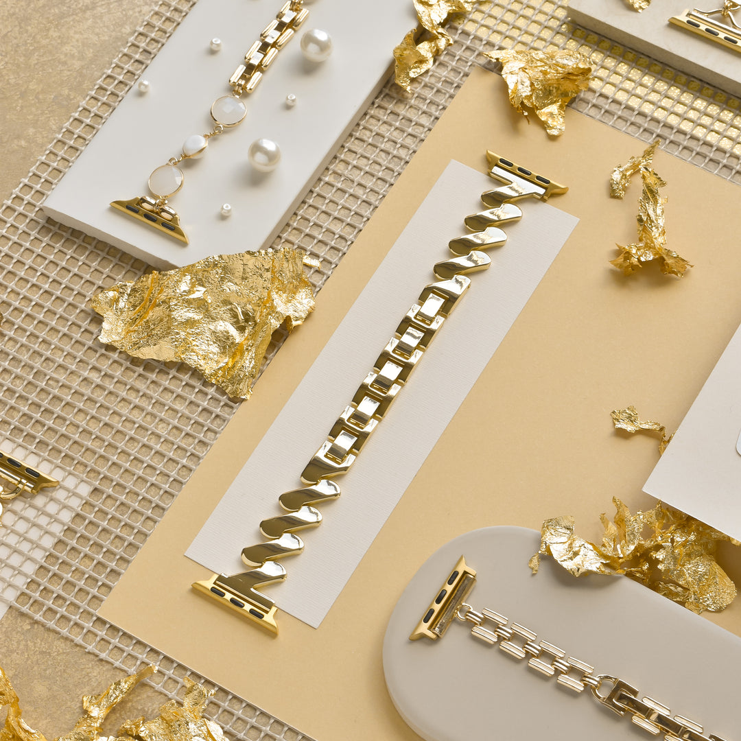 A flatlay showcases elegant gold and silver watch bands arranged on white and beige surfaces. The shiny bands, adorned with subtle details, are artistically placed among scattered pieces of gold leaf and a grid-patterned metallic backdrop. Featured prominently is the sleek Get Wavy Apple Watch Strap - Gold by Coconut Lane, designed for various Apple Watch models.