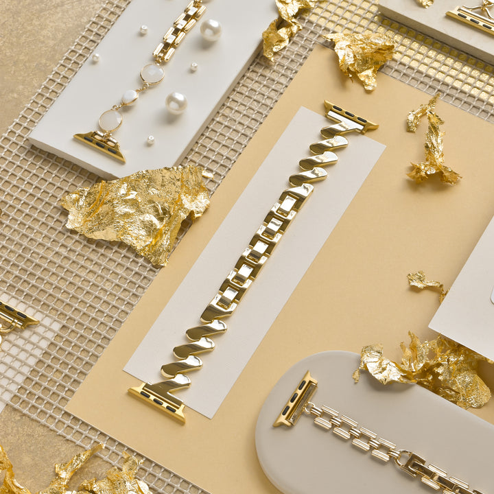 A flatlay showcases elegant gold and silver watch bands arranged on white and beige surfaces. The shiny bands, adorned with subtle details, are artistically placed among scattered pieces of gold leaf and a grid-patterned metallic backdrop. Featured prominently is the sleek Get Wavy Apple Watch Strap - Gold by Coconut Lane, designed for various Apple Watch models.