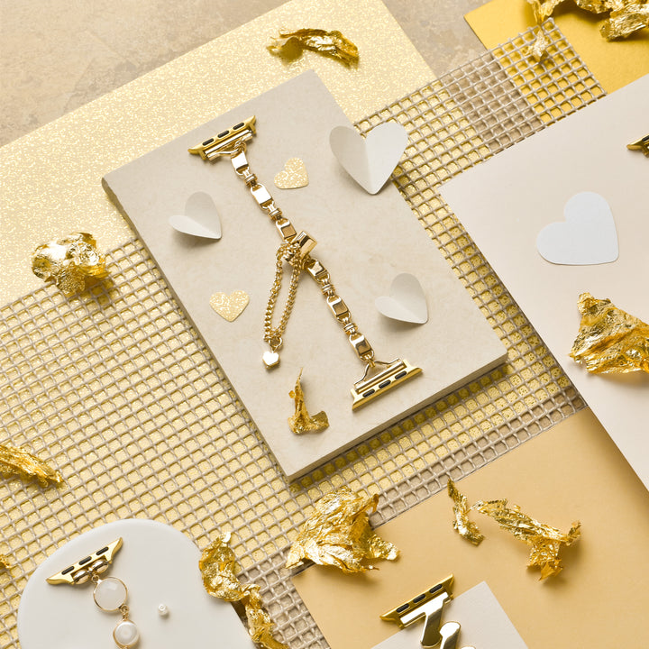 A stylish flatlay features gold jewelry including the Simple Gold Bracelet Apple Watch Strap by Coconut Lane, a textured surface, and scattered gold foil with white paper hearts. The arrangement also includes a stainless steel adjustable strap for an Apple Watch, along with simple gold rings and earrings. All are displayed on cream and gold backgrounds, creating a luxurious and elegant scene.
