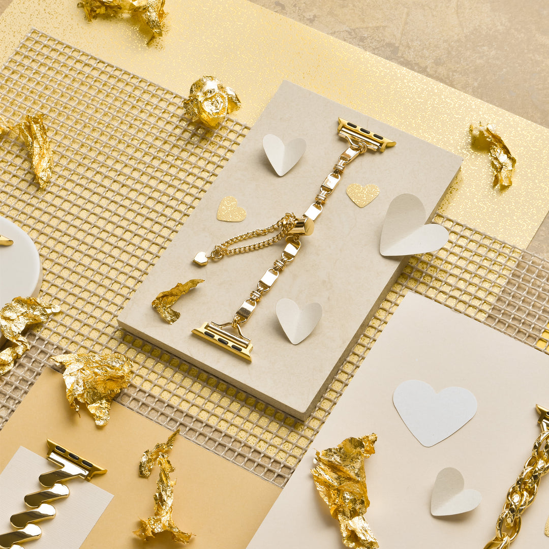 A Simple Gold Bracelet Apple Watch Strap by Coconut Lane is displayed on a beige surface, surrounded by heart-shaped cutouts and gold foil pieces. The scene is set against a gold and beige textured background, creating a luxurious and elegant aesthetic, reminiscent of its refined design.