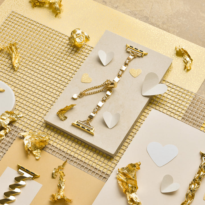A Simple Gold Bracelet Apple Watch Strap by Coconut Lane is displayed on a beige surface, surrounded by heart-shaped cutouts and gold foil pieces. The scene is set against a gold and beige textured background, creating a luxurious and elegant aesthetic, reminiscent of its refined design.