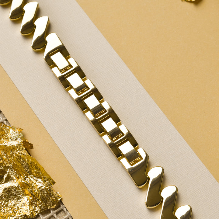 Close-up of a gold link chain bracelet placed diagonally on a beige and white background. The bracelet features rectangular links and shiny surfaces that reflect light, reminiscent of the luxe Get Wavy Apple Watch Strap - Gold by Coconut Lane. Fragments of gold foil are visible in the bottom left corner.