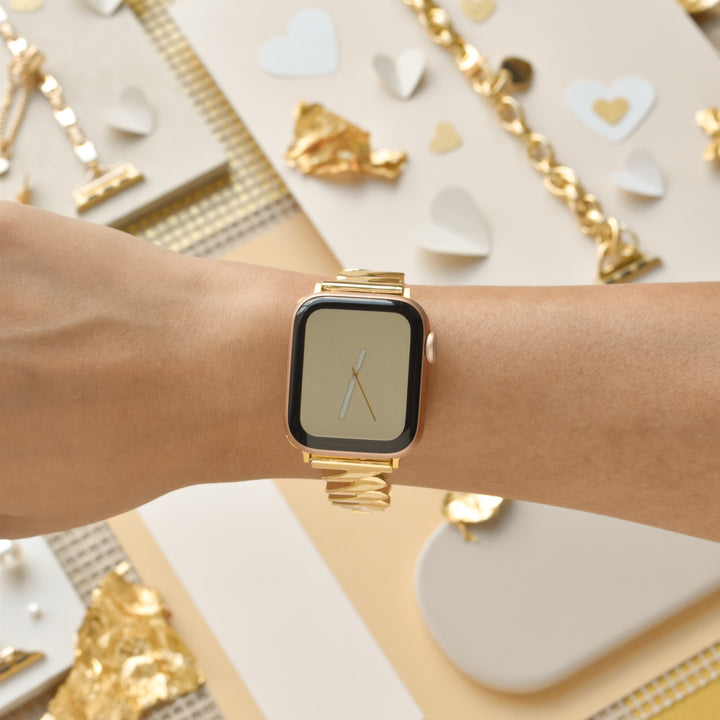 A left hand wearing a Get Wavy Apple Watch Strap in Gold by Coconut Lane, with a blank screen. The background features decorator paper hearts, gold foil, and a gold bracelet, arranged in a neutral, pastel setting. The image is focused on the stylish watch strap and the decorative backdrop, highlighting the elegance of this accessory.