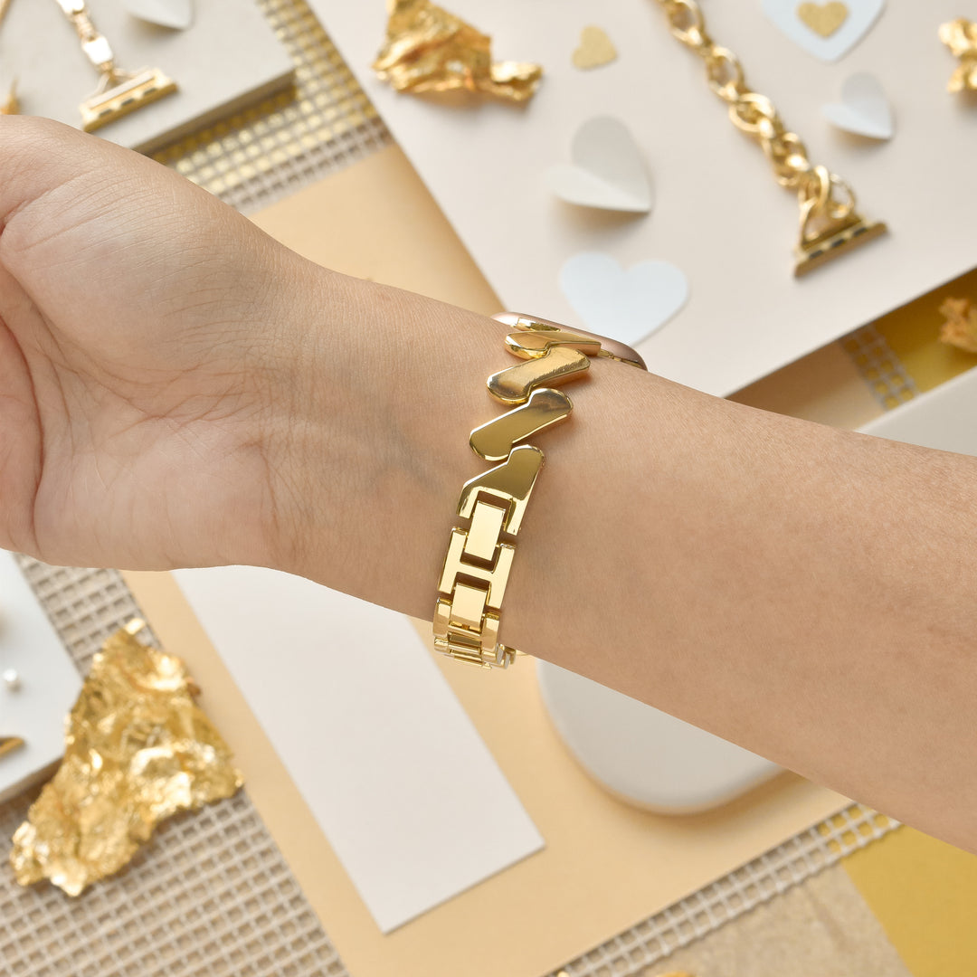 A wrist adorned with a gold bracelet featuring interlocking heart and rectangular links, complemented by the Get Wavy Apple Watch Strap - Gold from Coconut Lane. The background has materials used for jewelry making, such as gold foil and paper hearts, scattered on a beige and white surface.