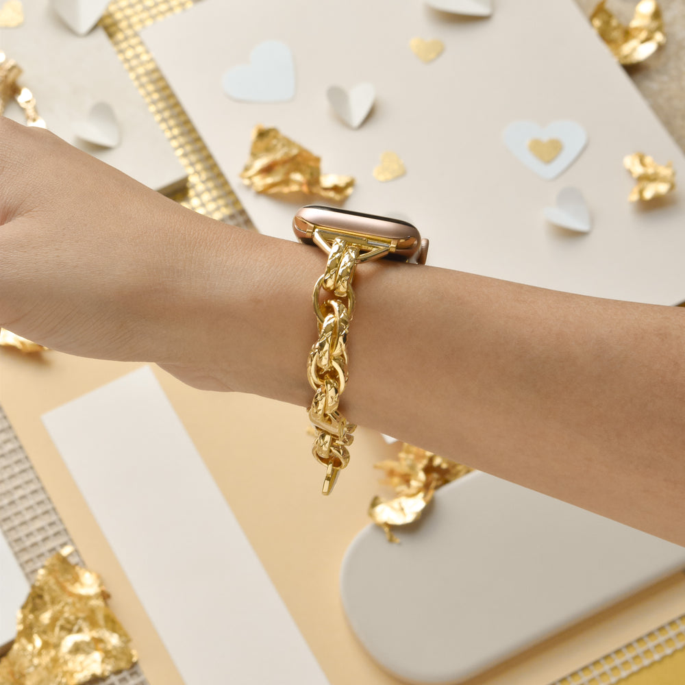 Close-up of a person’s wrist adorned with Coconut Lane's Chain Bracelet Apple Watch Strap in Gold. The background showcases a variety of craft materials, including white and gold paper hearts, gold foil scraps, and beige papers, forming a decorative and stylish scene.
