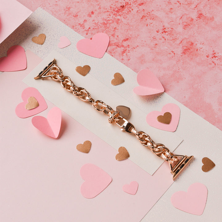 The Chain Bracelet Apple Watch Strap in Rose Gold by Coconut Lane is showcased on a light pink surface, adorned with small, scattered heart-shaped cutouts in shades of pink and gold. The backdrop features textured papers in pink and white, creating a romantic and elegant setting.