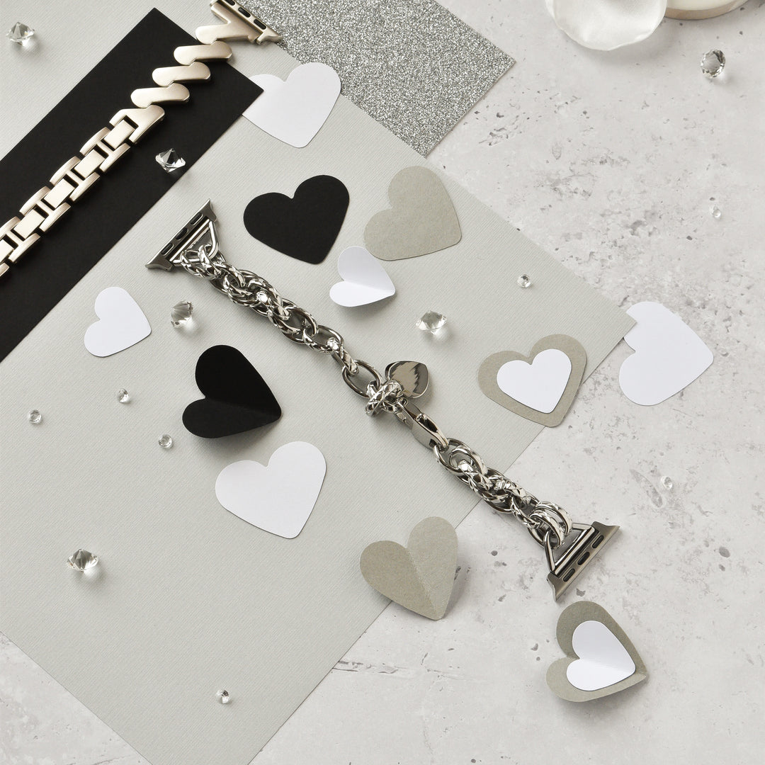 A Chain Bracelet Apple Watch Strap in silver by Coconut Lane lies on a textured surface surrounded by black, white, and gray paper hearts. Small clear gems are scattered around, adding a touch of sparkle to the arrangement. A cream-colored bracelet is visible at the top left of the image.