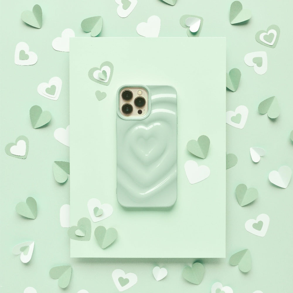 A smartphone with a Melting Heart Phone Case in Sage from Coconut Lane, featuring an extra protective raised heart design, is placed on a light green surface. Surrounding the phone are small paper hearts in various shades of green and white, creating a charming, romantic aesthetic.
