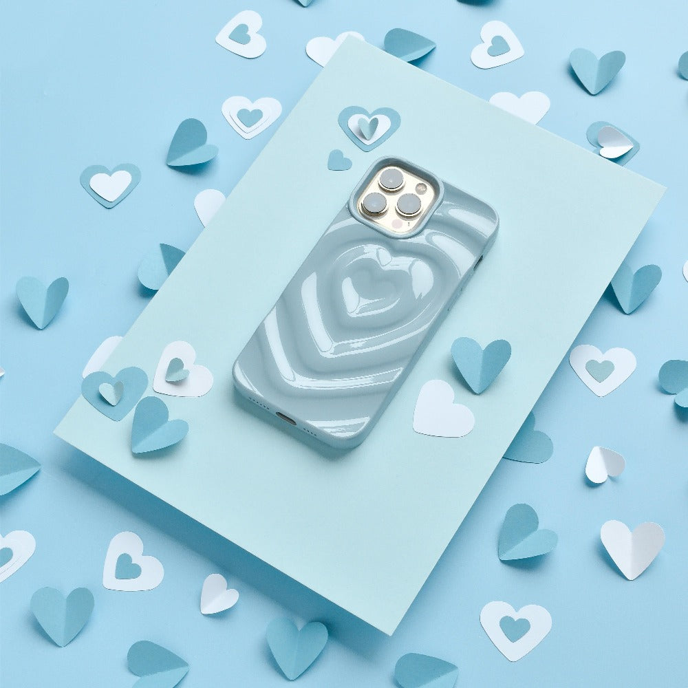 A smartphone with the Coconut Lane Melting Heart Phone Case in Baby Blue is placed on a light blue piece of paper. The background is decorated with scattered small heart-shaped cutouts in light blue and white.