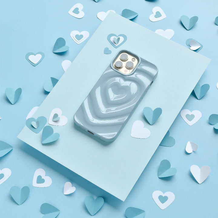 A smartphone with the Coconut Lane Melting Heart Phone Case in Baby Blue is placed on a light blue piece of paper. The background is decorated with scattered small heart-shaped cutouts in light blue and white.