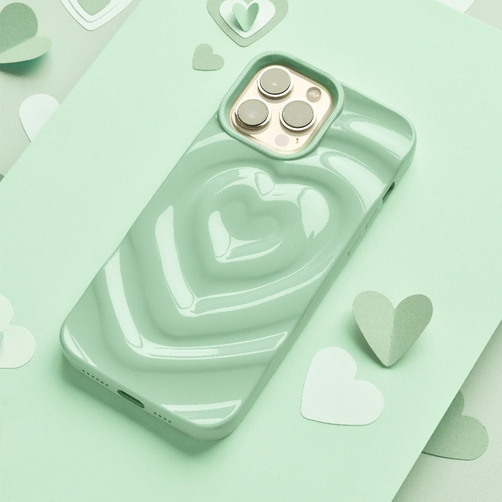 A sage-green smartphone with Coconut Lane's Melting Heart Phone Case rests on a matching mint green background. Small paper hearts in various shades of green are scattered around the phone, creating a cohesive and playful aesthetic.