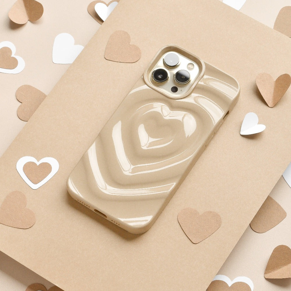 A biscuit beige iPhone with a glossy, extra protective Melting Heart Phone Case by Coconut Lane, featuring a 3D heart design, is placed on a beige surface. The background is decorated with scattered white and brown heart-shaped paper cutouts.