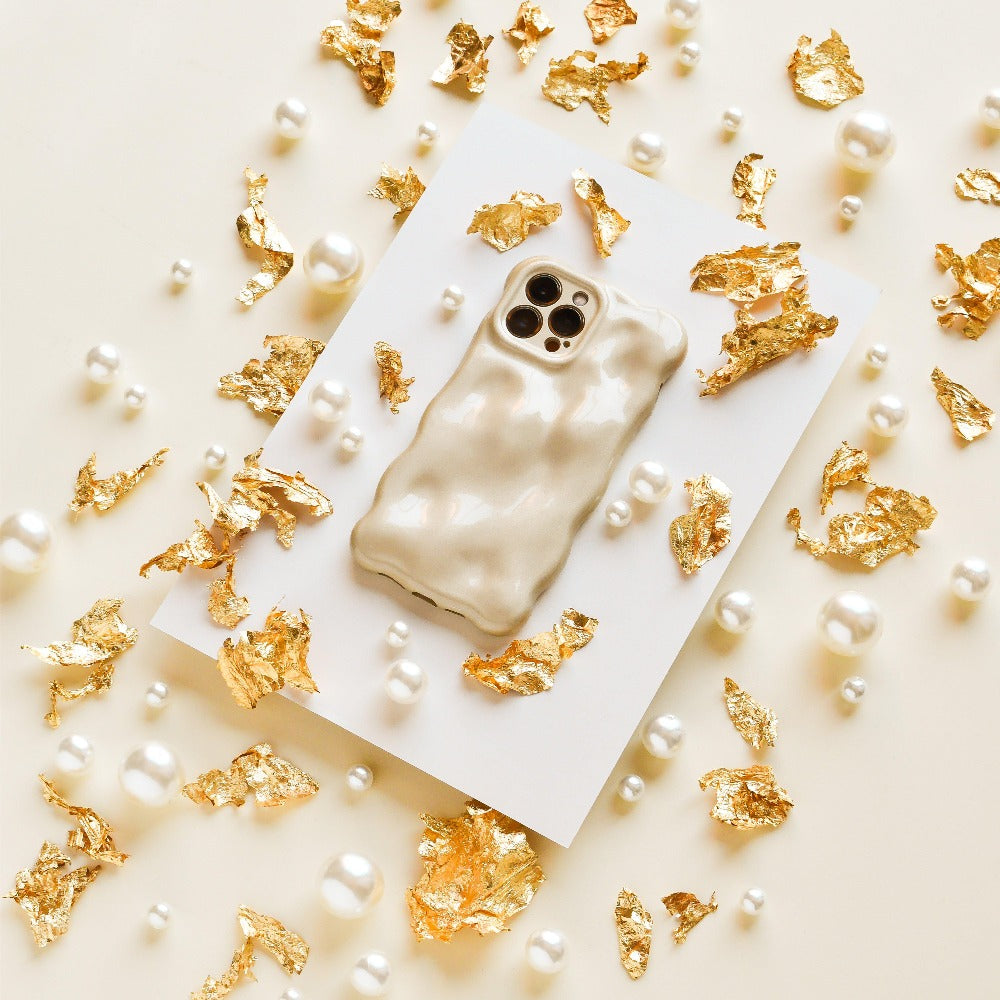 An iPhone15 adorned with Coconut Lane's 3D Pearl Phone Case - Gold rests on a white rectangular card. Scattered around are elegant gold foil pieces and 3D pearls against a beige background, crafting an extraordinarily luxurious composition.