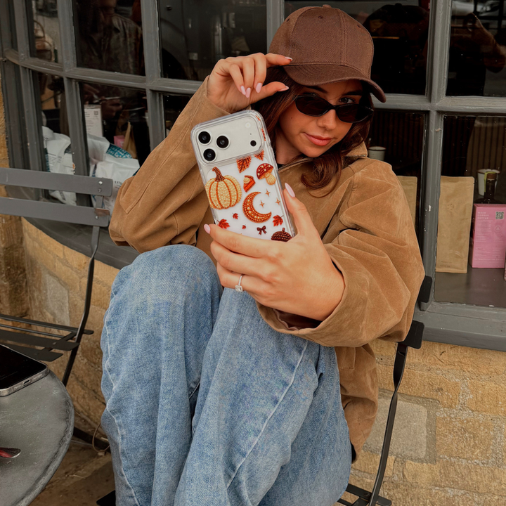 A woman wearing a brown cap, brown suede jacket, and blue jeans holds up a clear phone case decorated  with autumn-themed beaded effect designs including pumpkins,, mushrooms, leaves, shown in a lifestyle cafe setting. 