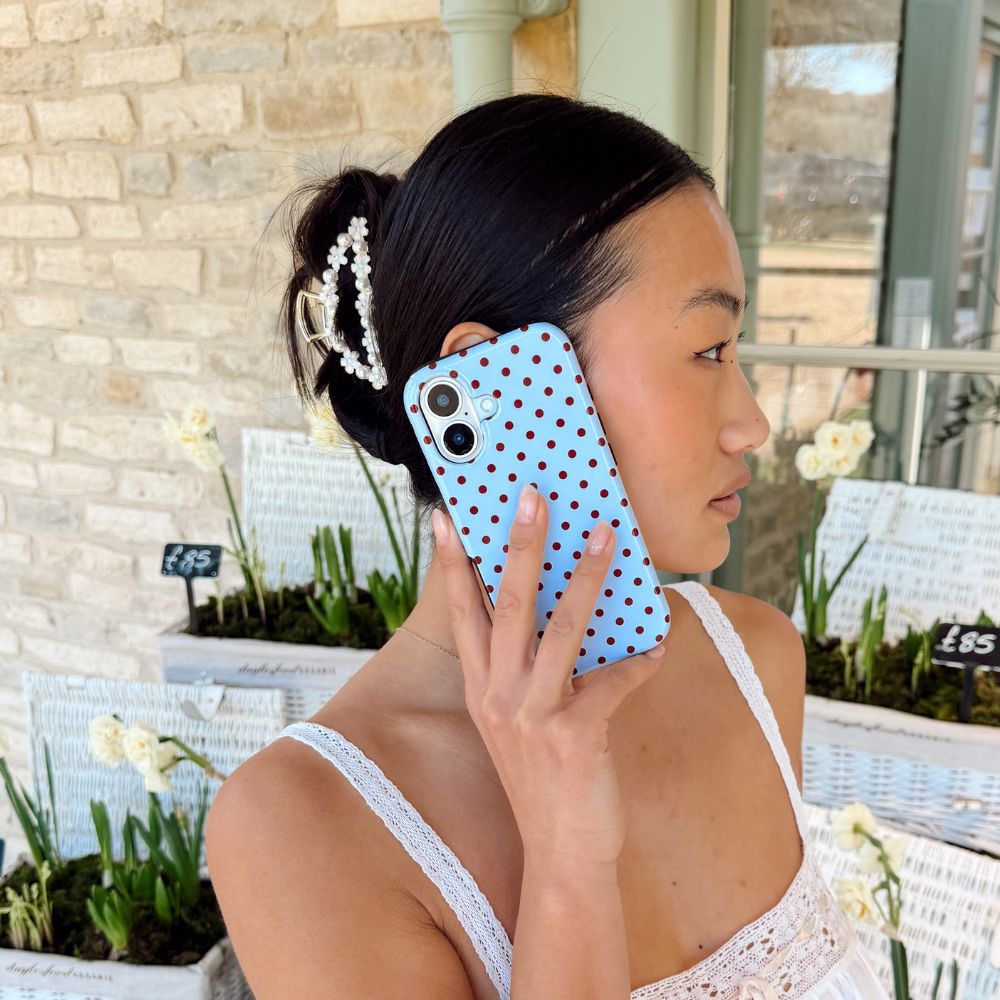 Woman wearing Coconut Lane Flowers & Pearls Claw Clip, holding a blue polka dot phone near flower boxes.