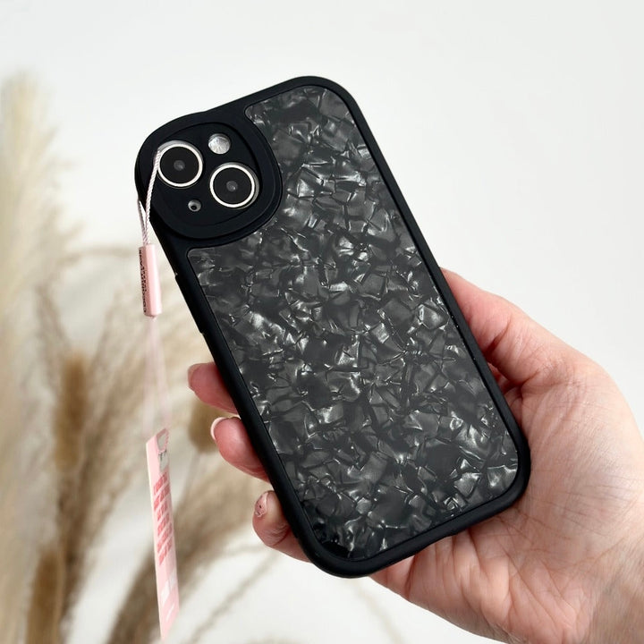 A hand holding a Moonstone Black Phone Case by Coconut Lane, featuring a textured, marble-like pattern with a small pink tag attached to it. The black phone case offers durable protection. In the background, there are blurred light-colored pampas grass.