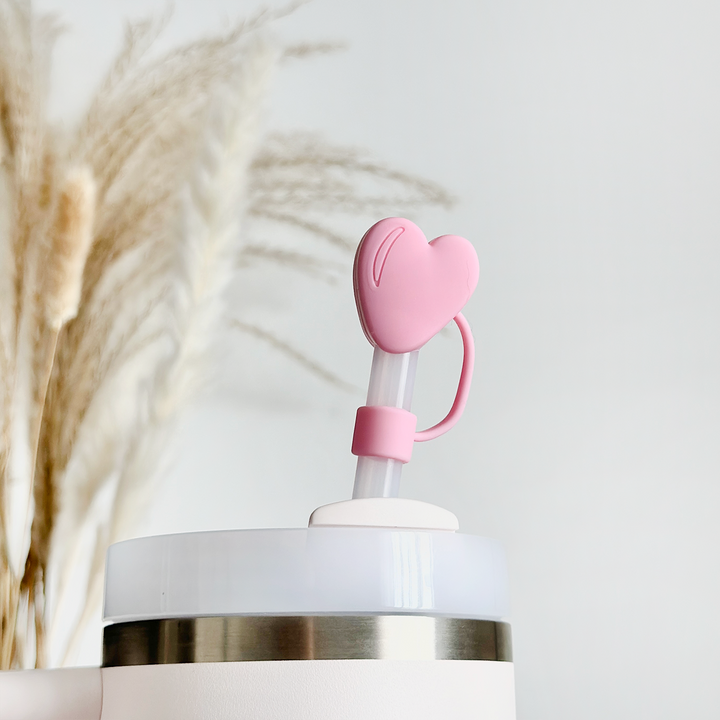 A white tumbler with a translucent straw is topped with Coconut Lane's Pink Heart Straw Cover, highlighting its flexible design. In the background, blurred pampas grass adds a touch of natural decor to the bright and minimalistic scene.