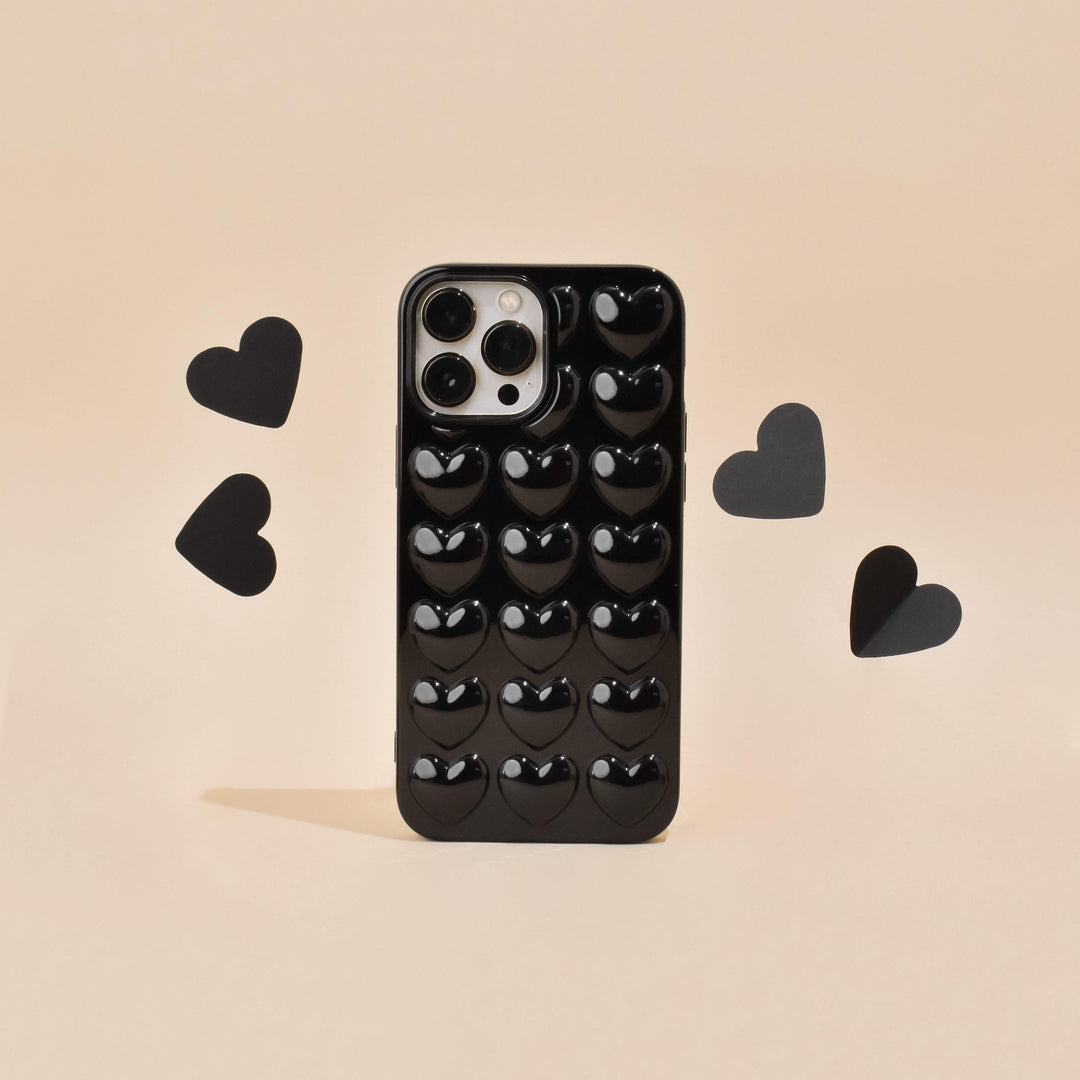 A "Coconut Lane" 3D Hearts Phone Case - Glossy Black is displayed against a tan background. Four black paper hearts surround the highly protective case, with two on each side, creating a playful and romantic theme. The case features a grid of glossy, raised heart shapes.