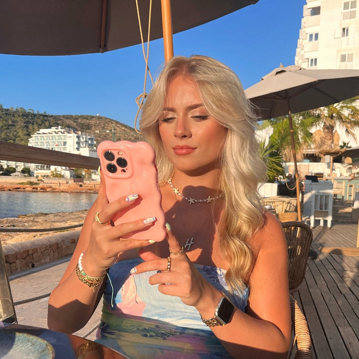 A blonde woman, seated at an outdoor café by the waterfront, holds a Coconut Lane 3D Gummy Phone Case - Pink shaped like a bear. Dressed in a chic and colorful floral outfit, she accessorizes with jewelry and an Apple Watch. The background features buildings, hills, and umbrellas under a clear blue sky.