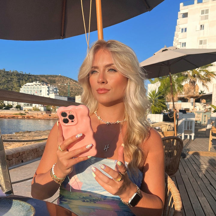 A woman with long blonde hair sits outside at a wooden table under an umbrella, holding a 3D Gummy Phone Case - Pink from Coconut Lane shaped like a bear. She is dressed in a light blue dress, has several rings on her fingers and painted nails. The background features buildings and palm trees.
