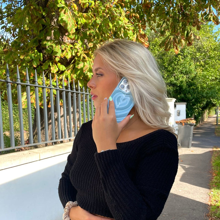 A woman with blonde hair, wearing a black long-sleeve top, holds a phone to her ear on a treelined sidewalk next to a fence. The sun is shining, casting shadows on her and her Coconut Lane Melting Heart Phone Case in baby blue featuring a textured design.